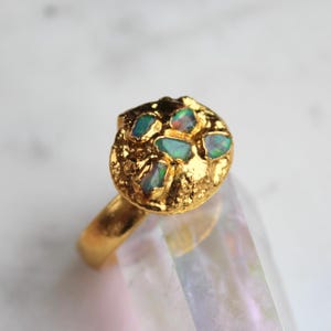 Puede incluir: Un anillo de oro con un engaste circular texturizado adornado con varias piedras de ópalo. Los ópalos muestran una mezcla de colores azules, verdes e iridiscentes. La banda del anillo es lisa y pulida, contrastando con el engaste texturizado.