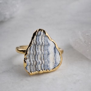 Può includere: Un anello color oro con una grande pietra piatta e irregolare al centro. La pietra presenta un motivo ondulato a strati nei toni del bianco, grigio e blu, con un bordo in metallo color oro lucido che incornicia l'intero perimetro.