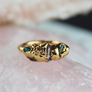 Op de afbeelding: Een goudkleurige ring met een getextureerde band en een cluster van ruwe edelstenen. De edelstenen omvatten een centrale heldere kristal en kleinere turquoise en blauwgroene stenen. De ring is geplaatst tegen een zachte roze achtergrond.