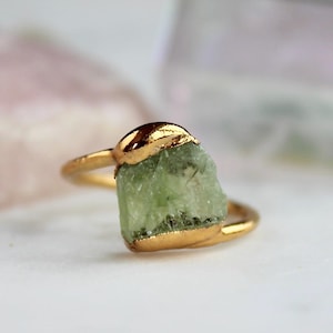 Könnte beinhalten: Ein goldfarbener Ring mit einem rohen, hellgrünen Edelstein. Der Edelstein hat eine unregelmäßige Form und ist teilweise in Gold gefasst. Der Ring ist dünn und ebenfalls goldfarben. Der Hintergrund ist verschwommen, mit weichen Rosa- und Weißtönen.