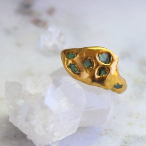May include: A gold-colored ring with an organic shape, featuring several emerald-green stones set into the metal. The ring is displayed on a white, crystalline surface, creating a contrast between the warm gold and cool, clear crystals.