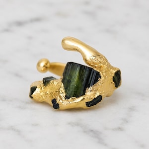 May include: A gold ring featuring a raw, dark green tourmaline crystal set within a textured, organic metal band. The design includes a prominent, vertically striated green stone surrounded by uneven, molten-style gold plating.
