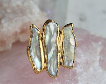 May include: A gold ring with three irregular, white, iridescent pearl-like stones. The stones are set in a gold-coloured metal. The ring is displayed on a pink surface. The design is unique and elegant.