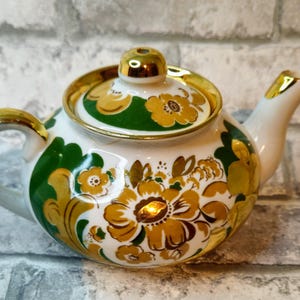 Könnte beinhalten: Eine kleine weiße Teekanne mit goldenem Deckel und Henkel. Die Teekanne ist mit einem floralen Muster in Grün- und Goldtönen verziert.