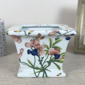 May include: White ceramic planter with a square shape and a floral design. The planter features pink, blue, and green flowers with a white background.