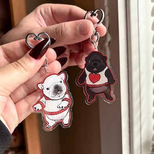 May include: Two keychains featuring cartoon bulldog designs. One keychain has a white bulldog wearing a red sweater, and the other has a black bulldog wearing a white shirt with a red heart. Both keychains have a silver heart-shaped clasp.