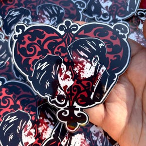 May include: Heart-shaped sticker featuring a couple in a passionate embrace, with blood splatters on their faces. The design includes a red filigree pattern and black outlines, creating a gothic aesthetic. The sticker is held in a hand.
