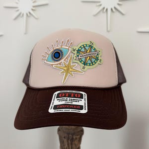 May include: A brown and tan trucker hat with a mesh back. The hat features embroidered patches of an eye, compass, and star. The text "Wanderlust" is on the compass patch. The hat has a snap closure and the brand name "OTTO" is visible.