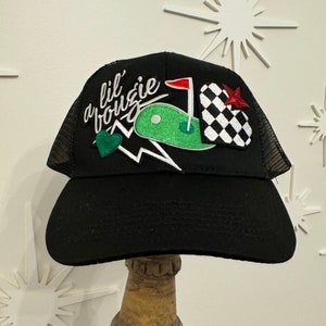May include: Black mesh trucker hat with a green golf course, red flag, and checkered heart patch. The hat also features a white lightning bolt and the text "a lil' bougie" in white cursive.