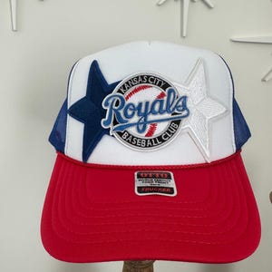 May include: A Kansas City Royals baseball cap with a red brim, white crown, and blue mesh back. The cap features a team logo with a baseball and star accents. The text "Royals" is displayed prominently.