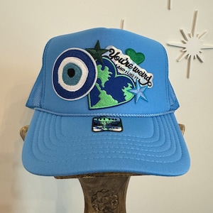 May include: A light blue trucker hat with a variety of patches, including a green heart with the text "You're Weird and I Like It", a blue evil eye, and a green and blue earth. The hat has a mesh back.
