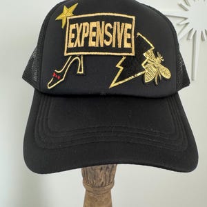 May include: Black trucker hat with a mesh back and a curved brim. The front features the word "EXPENSIVE" in gold embroidery, along with a gold star, a high heel, and a bee.