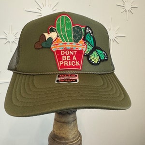 May include: A green trucker hat with a cactus and butterfly patch. The patch says "Don't Be A Prick".