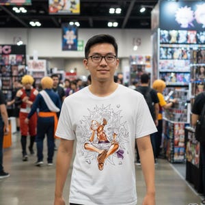 May include: A white t-shirt featuring a colorful anime character design. The graphic depicts a seated figure with a dynamic energy effect. The person wearing the shirt is in a crowded convention setting.