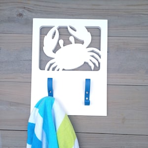 Puede incluir: Un gancho de pared de madera blanco con un recorte de un cangrejo y dos ganchos de metal azul. Una toalla a rayas azul y verde está colgada en uno de los ganchos.