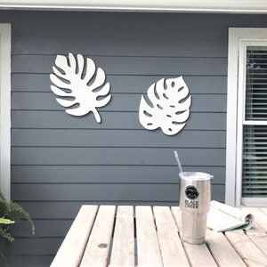 Puede incluir: Dos piezas de decoración de pared de hojas de monstera de madera blanca están montadas en una pared exterior de revestimiento gris. Un vaso plateado con un logotipo negro que dice "Black Creek" está sobre una mesa de madera.