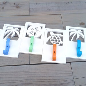Puede incluir: Seis ganchos de madera blancos con diferentes recortes de temática playera. Los ganchos están pintados en varios colores, incluyendo azul, verde, naranja y gris.
