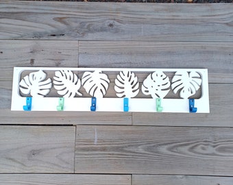 May include: White wooden wall-mounted coat rack with a cutout design of tropical leaves. The rack has six hooks with blue and green painted accents.