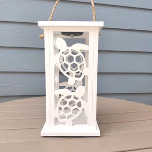 May include: White wooden lantern with cut-out sea turtle designs. The lantern has a rope handle and is designed to hold a light. The lantern is sitting on a brown surface.