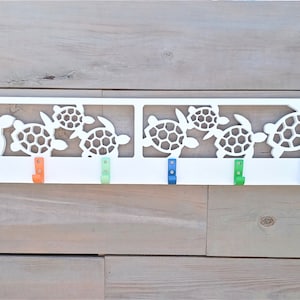 May include: White wooden wall mounted coat rack with a cutout design of sea turtles. The rack has six hooks in various colors: blue, orange, green, and light blue.