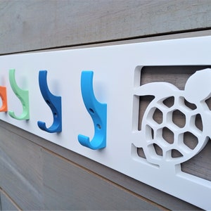 May include: White wooden wall-mounted coat rack with five hooks in different colors: blue, light blue, orange, green, and dark blue. The rack features a cutout of a turtle with a shell design.