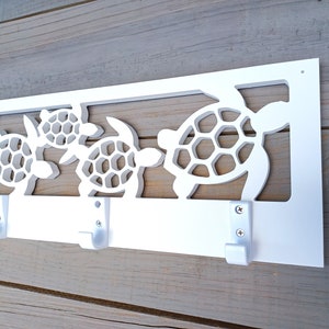 May include: White wooden wall mounted coat rack with three hooks. The rack is decorated with a cutout design of three sea turtles.