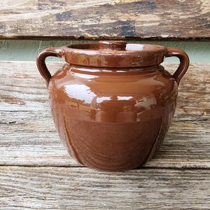 May include: A brown ceramic jar with a lid and two handles. The jar has a glossy finish and sits on a wooden surface. The jar is a traditional kitchenware item.