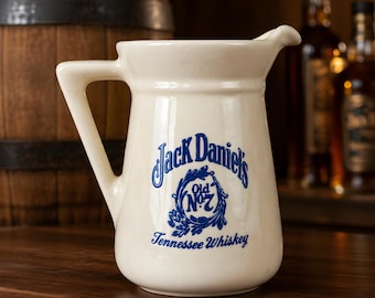 Puede incluir: Jarra de cerámica color crema con asa y boquilla. La parte delantera presenta el logotipo azul "Jack Daniel's Old No. 7 Tennessee Whiskey". La jarra está sobre una superficie de madera, con un barril de madera y botellas en el fondo.