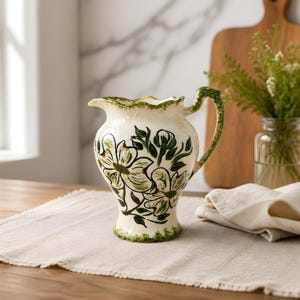 May include: Cream-colored ceramic pitcher with a green floral design. The pitcher has a scalloped rim and handle, with green accents. The floral pattern includes green leaves and white flowers. The pitcher is on a beige linen runner.