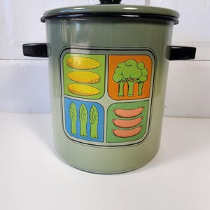 May include: A vintage green enamel pot with a black lid and handles. The pot features a colorful graphic of vegetables, including corn, broccoli, asparagus, and sausages. A retro kitchenware item.