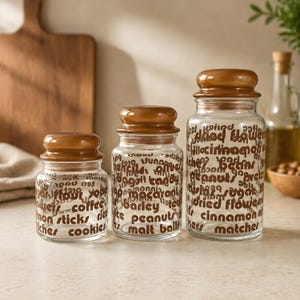 May include: Three clear glass jars with brown lids, each labeled with food-related words in a repeating pattern. The jars vary in size, with the largest jar containing words like "beans" and "cinnamon."