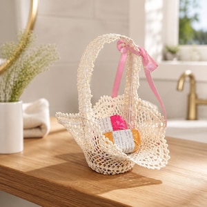 May include: A cream-colored crocheted basket with a scalloped edge and a single handle adorned with a light pink satin ribbon. Inside the basket are two small rectangular bars wrapped in paper, one with a pink floral pattern and one with orange.