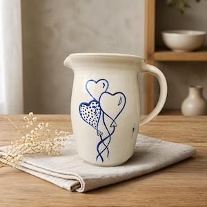 May include: Cream-colored ceramic pitcher with a blue heart balloon design. The pitcher has a handle and a spout for pouring. The heart balloons are connected by blue lines. The pitcher is sitting on a neutral-colored cloth.