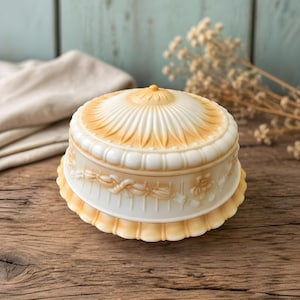 May include: A two-toned, antique-style cake stand with a domed lid. The lid and base are a creamy white with a golden-yellow trim. The base features a floral and ribbon design. The stand is on a wooden surface.