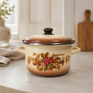 May include: Cream-colored enamel pot with a lid and side handles. The pot features a floral design with red berries, brown leaves, and yellow accents. The lid is brown with a dark brown knob. A classic kitchenware item.