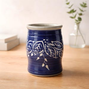 May include: A handcrafted, blue ceramic vase with a white leaf design. The vase has a cylindrical shape with a slightly flared base and a gray rim. The leaf pattern is repeated around the vase.