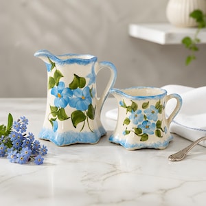 May include: Two ceramic pitchers with blue floral designs. The pitchers are white with blue trim and handles. The larger pitcher is approximately 7 inches tall, and the smaller pitcher is about 4 inches tall. Green leaves and blue flowers decorate the pitchers.