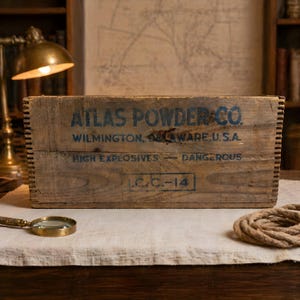 May include: A weathered wooden crate with the text "ATLAS POWDER CO." in blue lettering. The crate also displays "WILMINGTON, DELAWARE, U.S.A.", "HIGH EXPLOSIVES - DANGEROUS", and "C.C.-14". The crate is sitting on a white cloth.