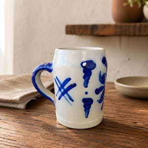 May include: A white ceramic mug with a blue handle and blue abstract designs. The mug has a cylindrical shape and a wide opening. The blue designs include cross-like shapes and stylized floral patterns. The mug sits on a wooden surface.