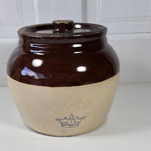 May include: A vintage stoneware crock with a dark brown glazed top and a tan base. The crock has a lid and a blue crown emblem with the number 3. This antique kitchenware is a classic piece of rustic decor.