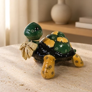 May include: A ceramic turtle figurine with a green head and shell, accented with yellow and black details. The turtle has a bow tie and is decorated with small white dots. The figurine is a decorative item.