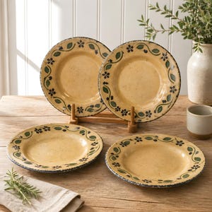 May include: A set of four ceramic plates with a rustic design. The plates have a yellow center, a blue rim, and are decorated with green leaves and blue flowers. Two plates are displayed on wooden stands.