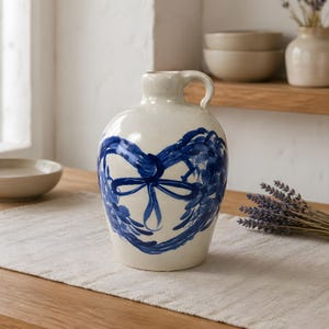 May include: Cream-colored ceramic jug with a blue heart-shaped design. The jug has a handle and a narrow neck. The heart design is painted with brushstrokes and a bow. The jug is sitting on a wooden table.