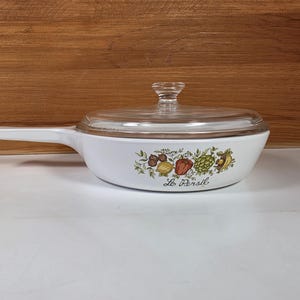 May include: White casserole dish with a clear glass lid and handle. The dish features a colorful floral and vegetable design with the text "Le Persil".