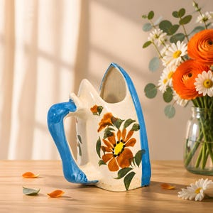 May include: A ceramic pitcher shaped like a vintage clothing iron, featuring a cream-colored body with hand-painted orange flowers, green leaves, and a bright blue handle and trim. The vessel has an open top and a sturdy, curved handle.