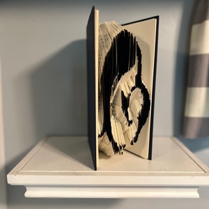May include: A book art sculpture of a black and white animal, possibly a bear, made by folding the pages of a book. The sculpture is displayed on a white shelf.