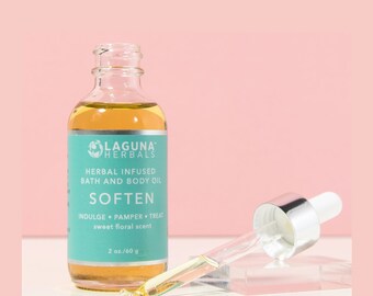 May include: A clear glass bottle containing a light yellow liquid. The bottle is labelled "LAGUNA HERBALS" and "HERBAL INFUSED BATH AND BODY OIL SOFTEN INDULGE PAMPER TREAT sweet floral scent 2 oz./60 g".