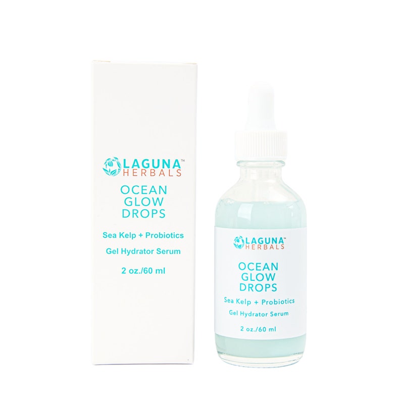 May include: A clear glass bottle with a blue liquid inside. The bottle has a white dropper top and a white label with the text "LAGUNA HERBALS OCEAN GLOW DROPS Sea Kelp + Probiotics Gel Hydrator Serum 2 oz./60 ml". The bottle is sitting on a white surface.