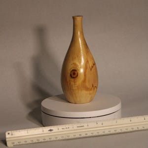 May include: A small, light brown wooden vase with a natural wood grain pattern. The vase has a narrow neck and a rounded body.