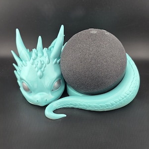 May include: A teal dragon-shaped speaker holder with a black speaker. The dragon has detailed features, including horns, eyes, and a curled tail. The speaker is round and covered in a dark gray fabric. The holder is designed to cradle the speaker.